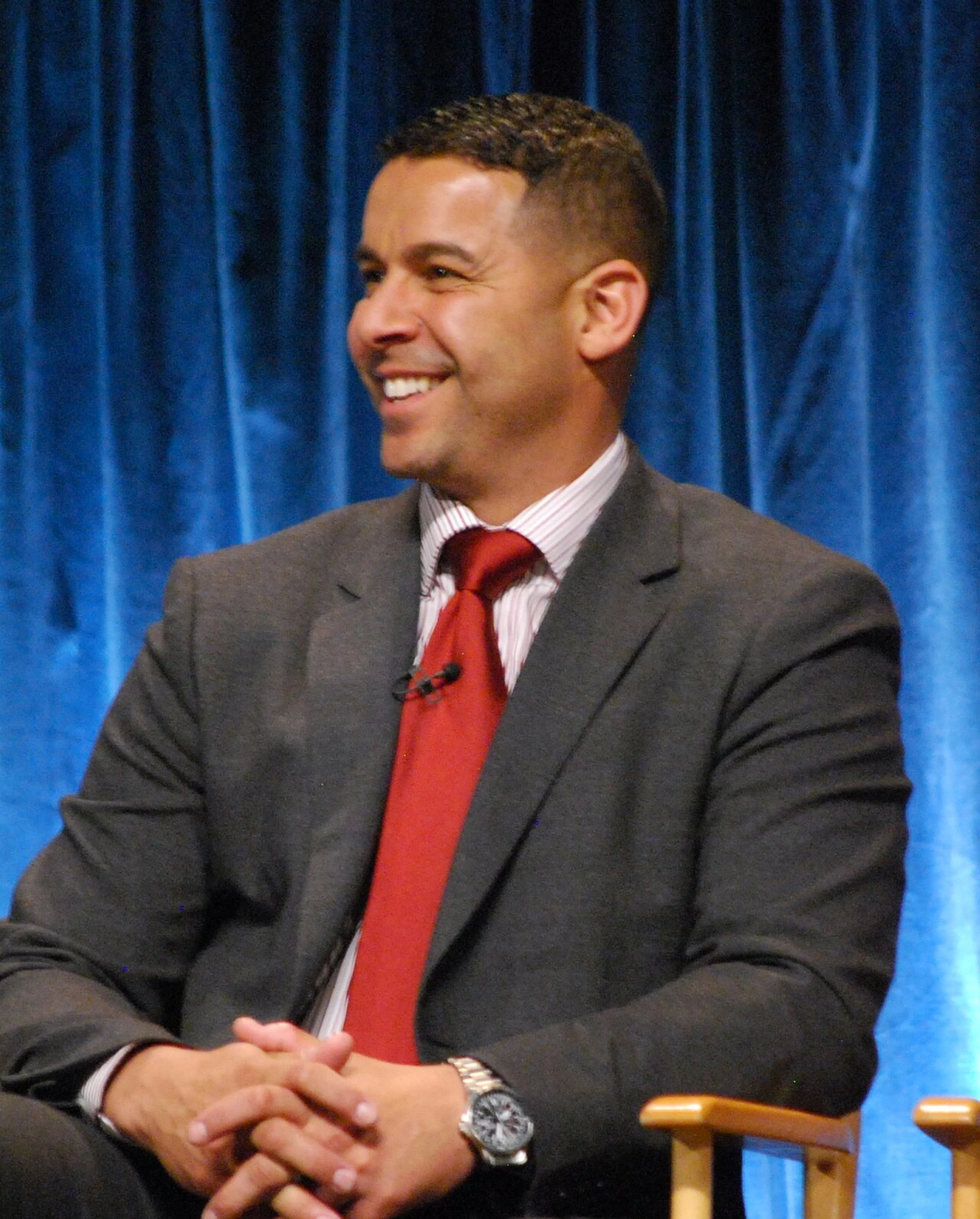 Jon Huertas seated on stage at PaleyFest, wearing a suit and red tie against a blue curtain background.