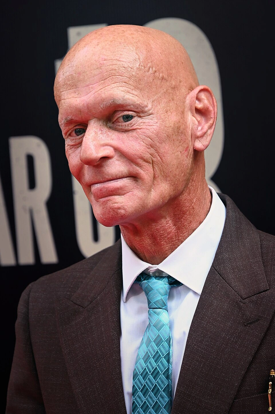 Portrait of Nick Yarris, bald man in a suit and turquoise tie at an event backdrop