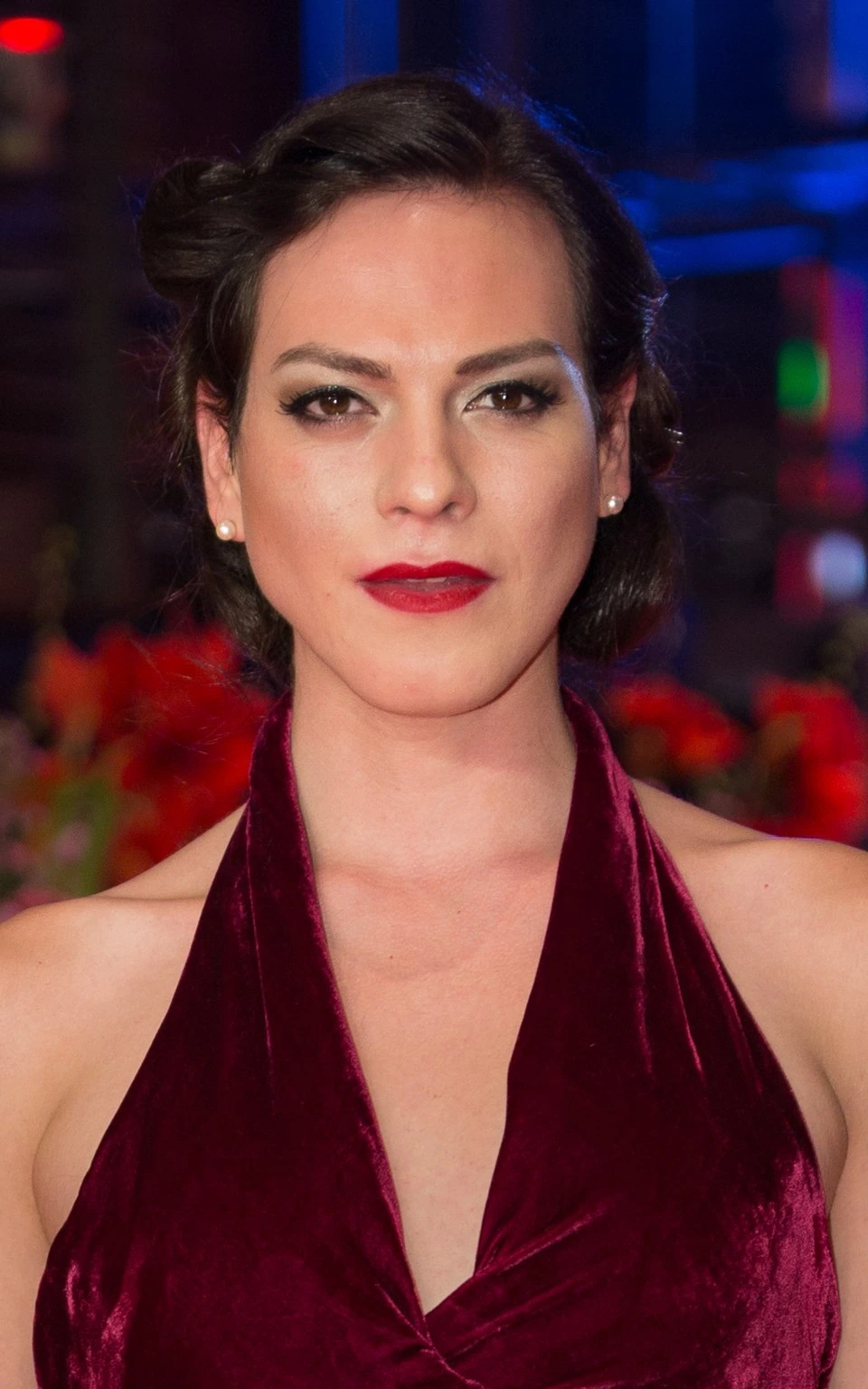 Portrait photo of Zuria Vega at an event, wearing a burgundy halter dress with red lipstick against a colorful blurred b