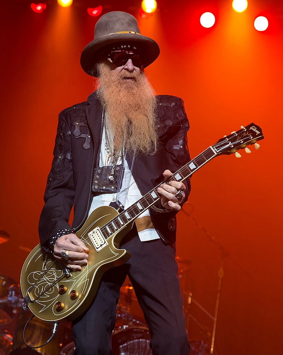 Billy Gibbons performing on stage with ZZ Top, playing guitar in front of the drum kit