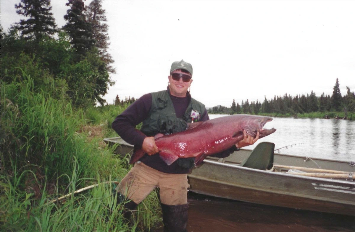 Billy Molls standing outdoors by a river holding a large fish near a boat