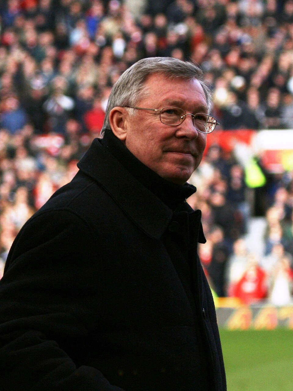 Sir Alex Ferguson photographed at a football stadium, wearing a dark coat and glasses, with a blurred crowd in the backg