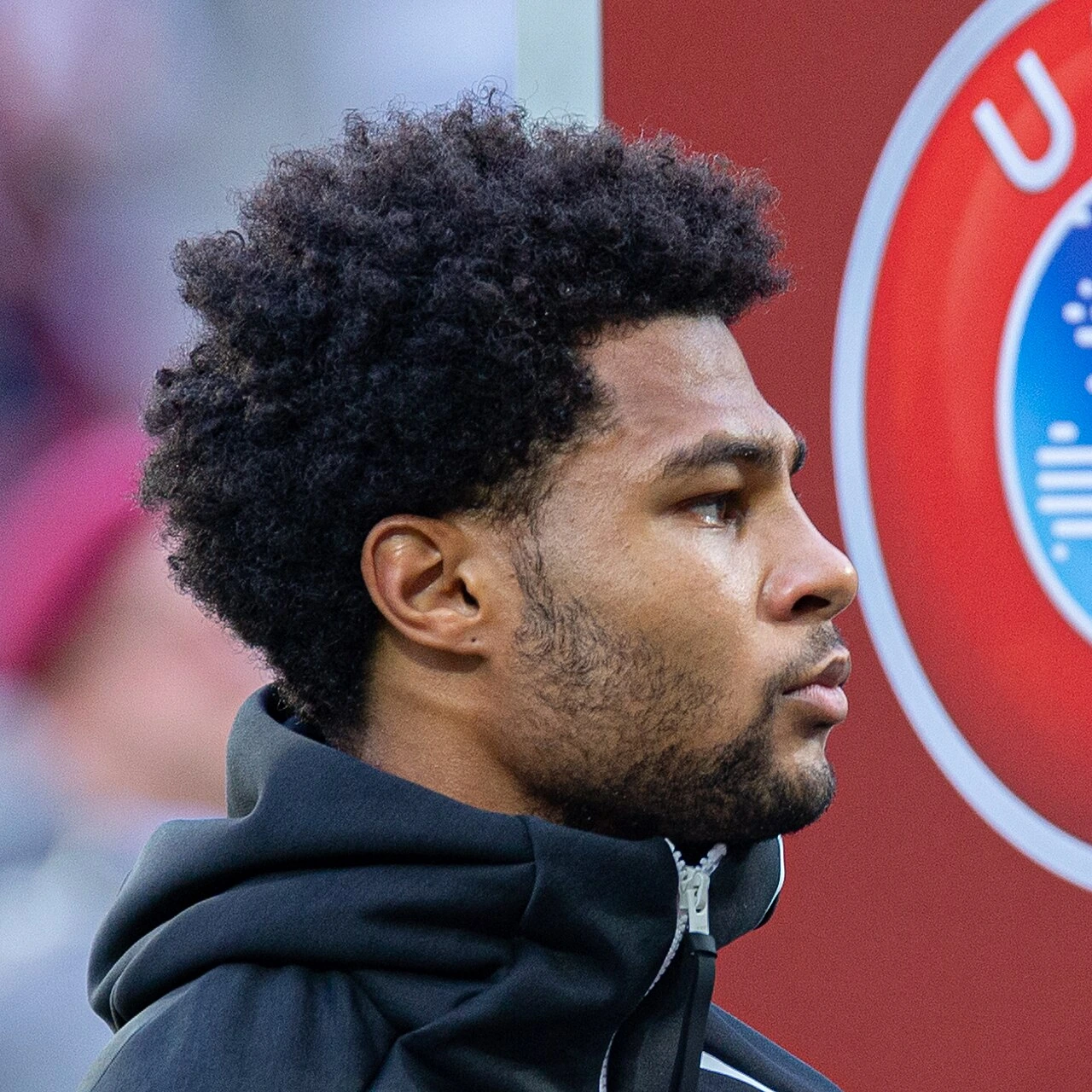 Serge Gnabry in profile during a match, wearing a dark jacket with a Bayern Munich backdrop behind him.