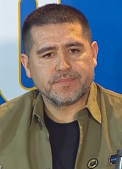 Juan Román Riquelme in a close-up portrait at an event, wearing a dark shirt and olive jacket against a blue background.