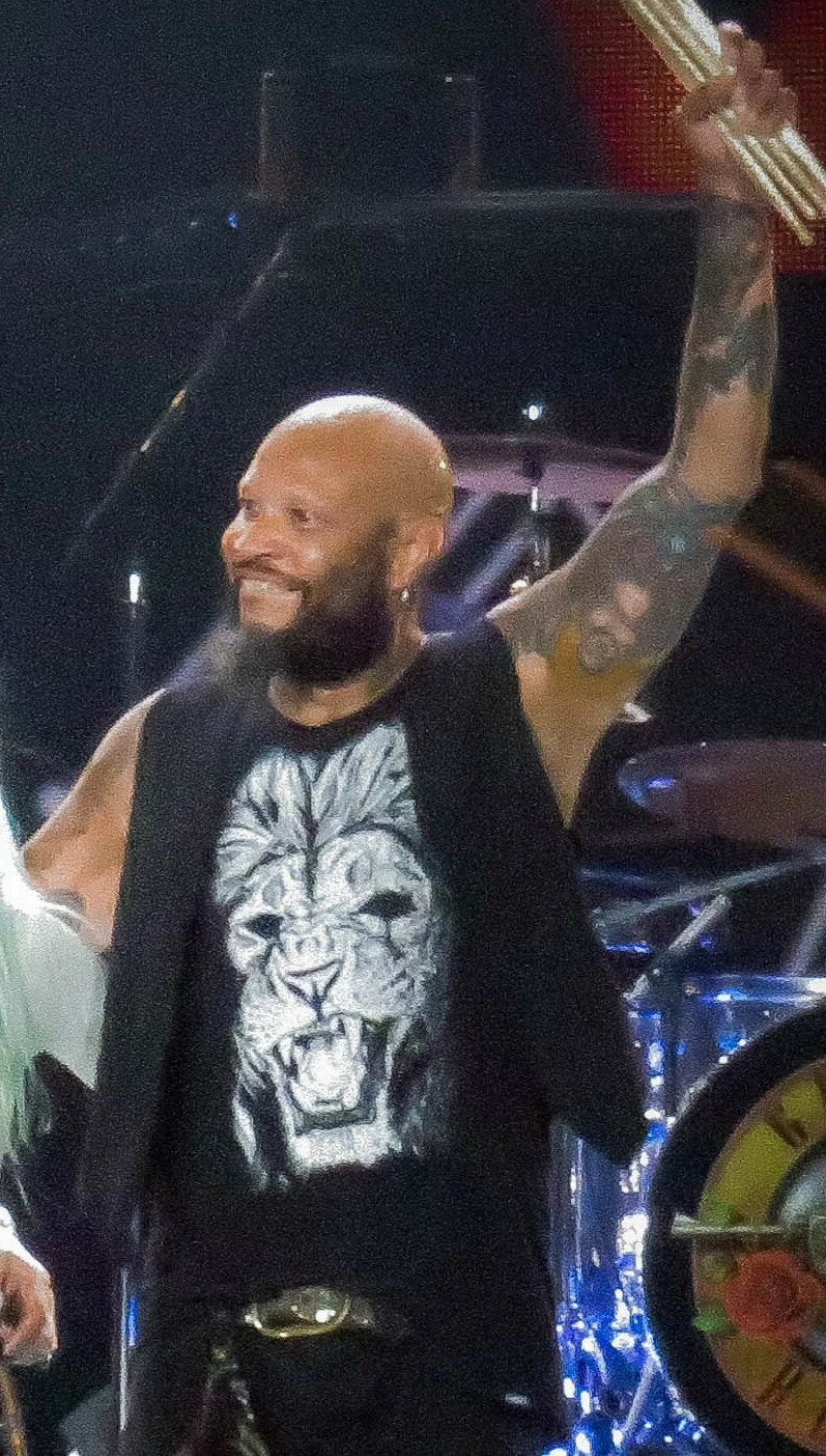 Frank Ferrer performing onstage during a Guns N' Roses concert, raising a drumstick and smiling under stage lights.