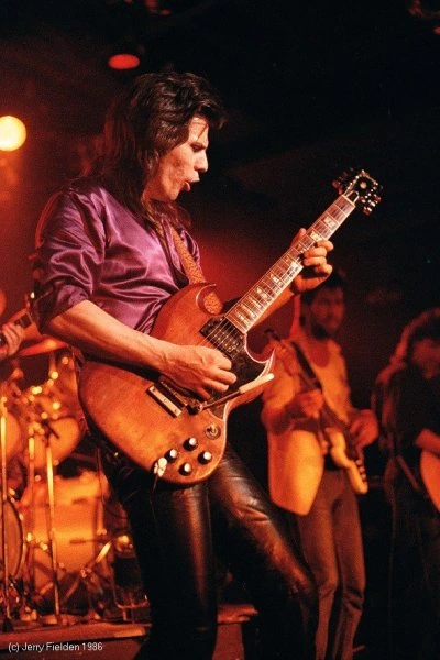 Frank Marino performing onstage with a guitar under warm stage lighting.