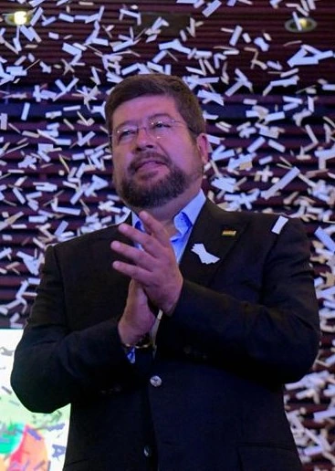 Samuel Doria Medina speaking at an event, wearing a dark suit and clapping
