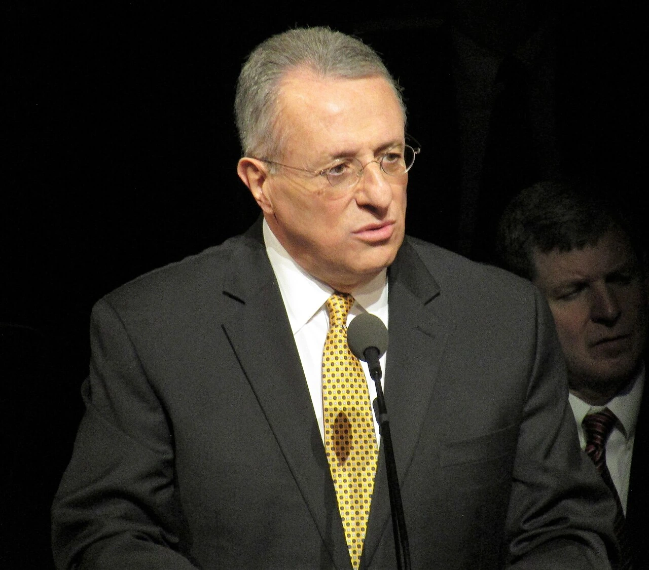 Ulisses Soares speaking at a podium in a dark setting, wearing a suit and yellow tie