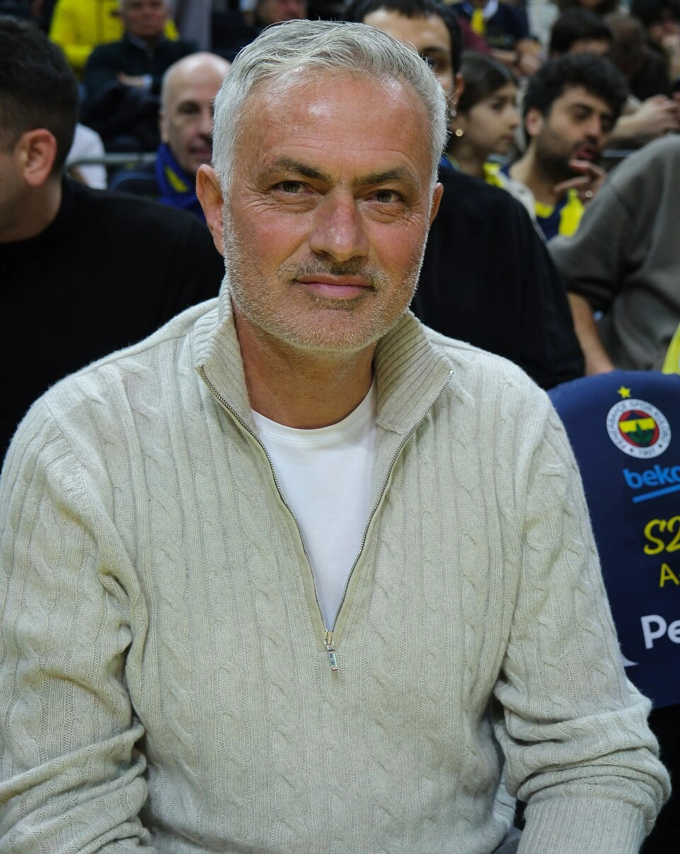 José Mourinho seated in a crowd at a football event, wearing a light beige zip-up sweater.