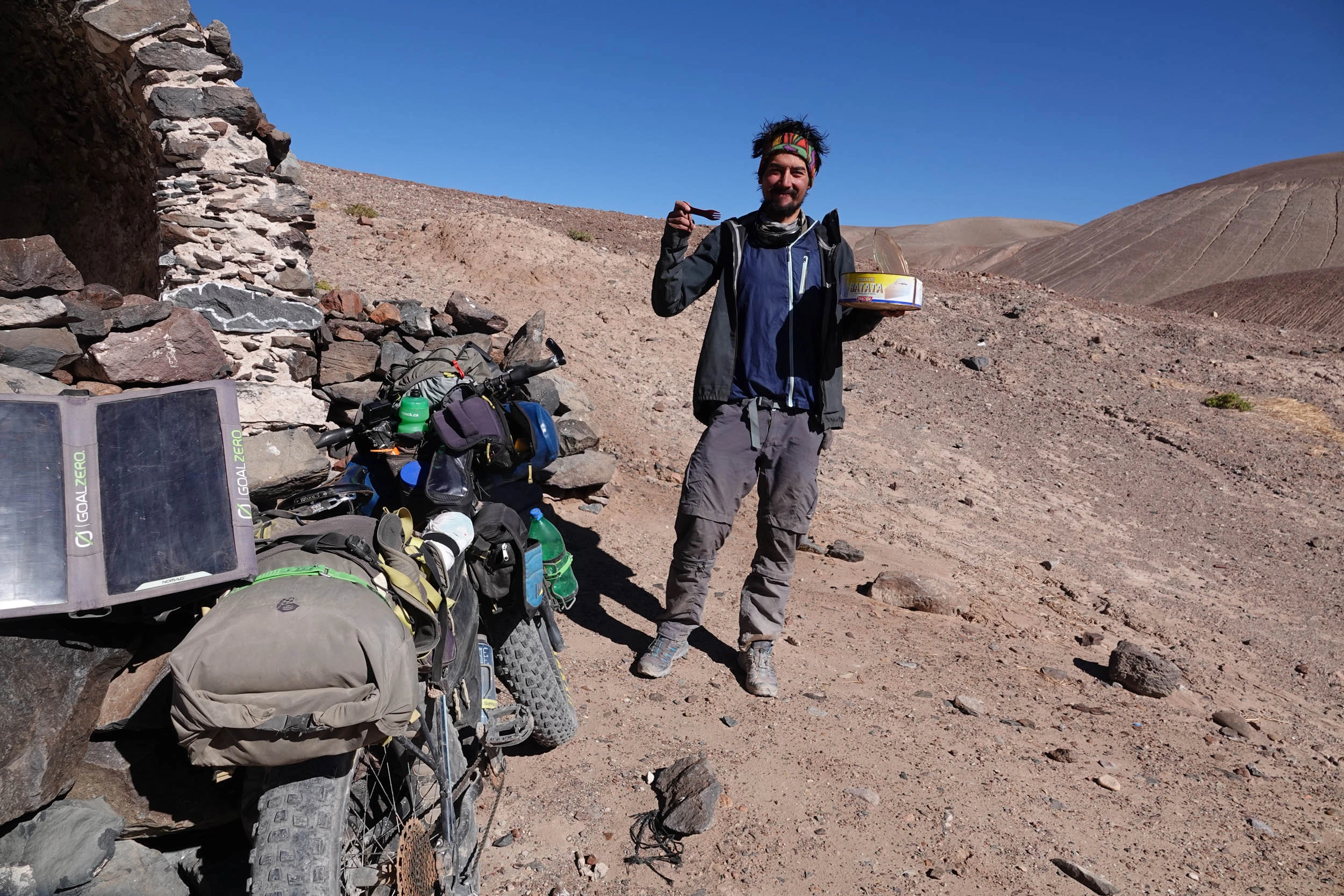 Iohan Gueorguiev standing beside a heavily loaded bike in a barren desert landscape under a clear blue sky.