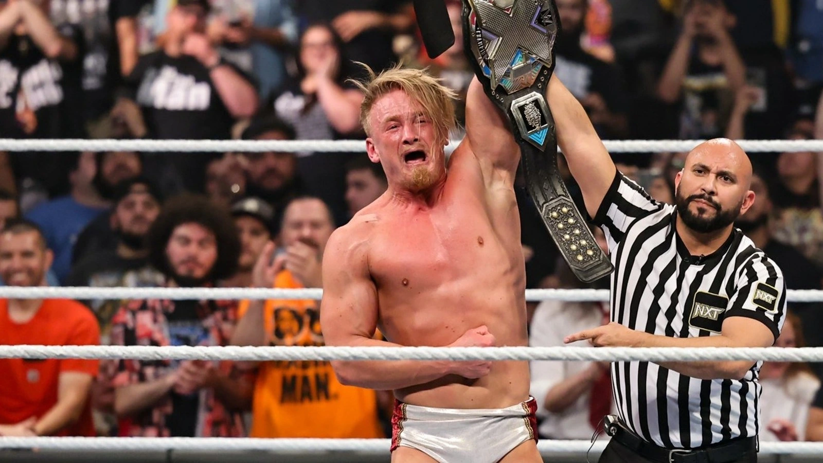 Ilja Dragunov wrestling in the ring during a match, shirtless and celebrating with a championship belt raised overhead.