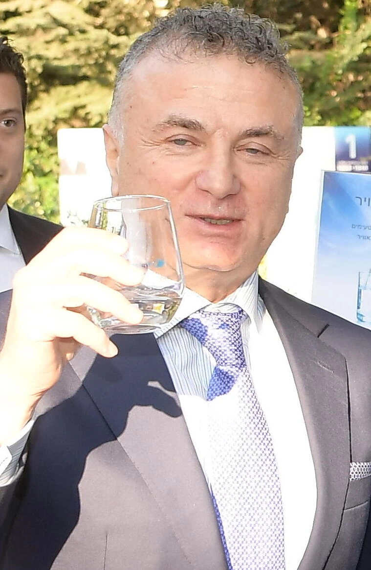 Mikhael Mirilashvili at an outdoor event, holding a glass while wearing a suit and tie.