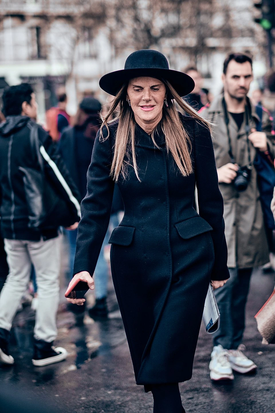 Anna Dello Russo walking in a black coat and wide-brim hat at Paris Fashion Week, photographed outdoors among a crowd.