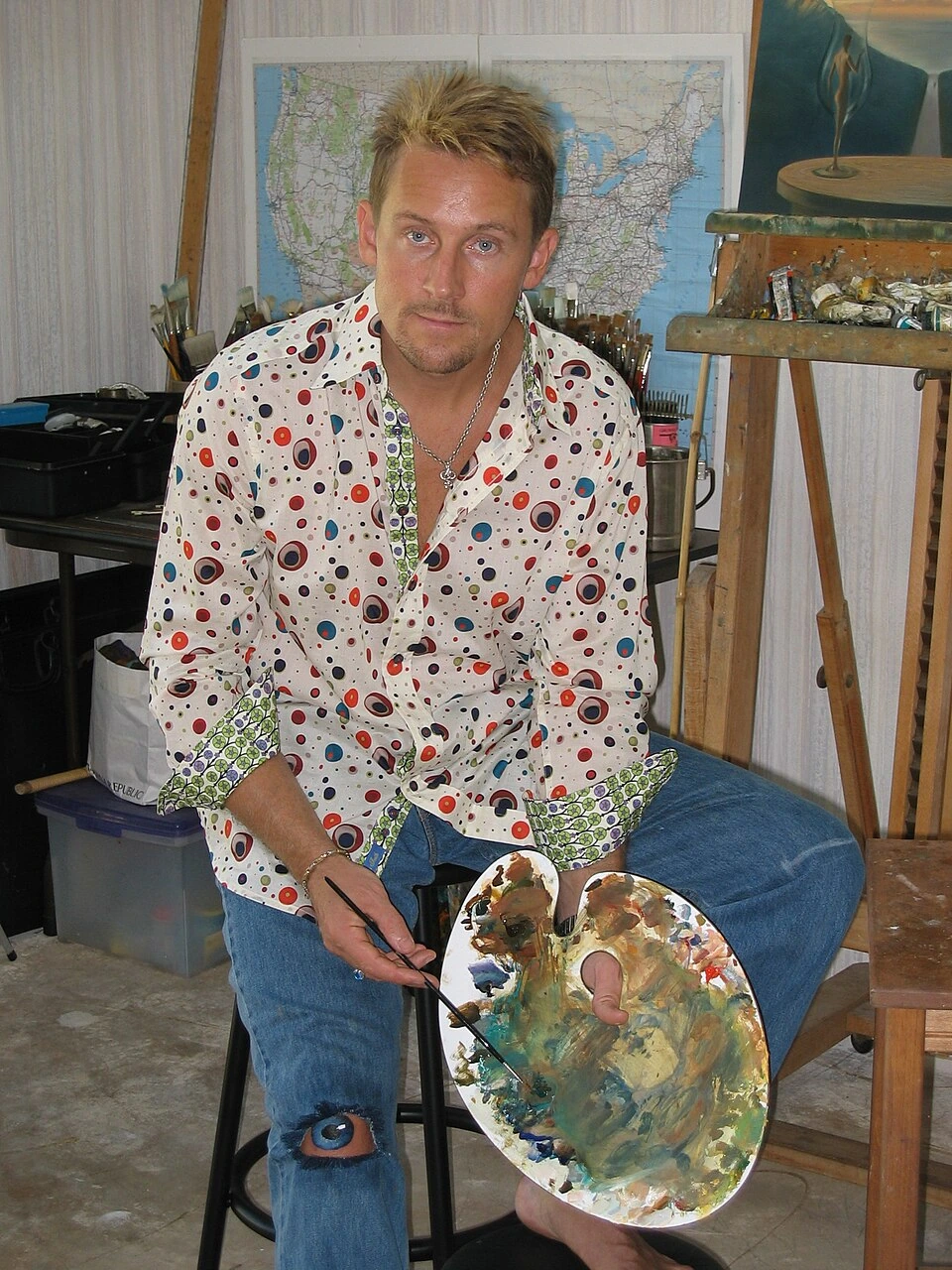 Vladimir Kush seated in his studio holding a paint palette, with easel and artwork in the background.