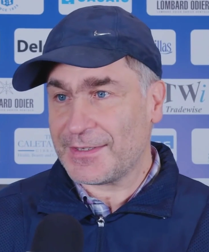 Vassily Ivanchuk at the chessboard during a tournament, wearing a dark jacket and cap, with a sponsor backdrop behind hi