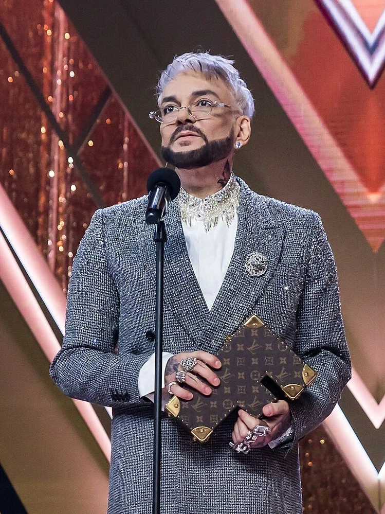 Philipp Kirkorov speaking on stage in a formal suit at an awards event, holding an item in front of a microphone.