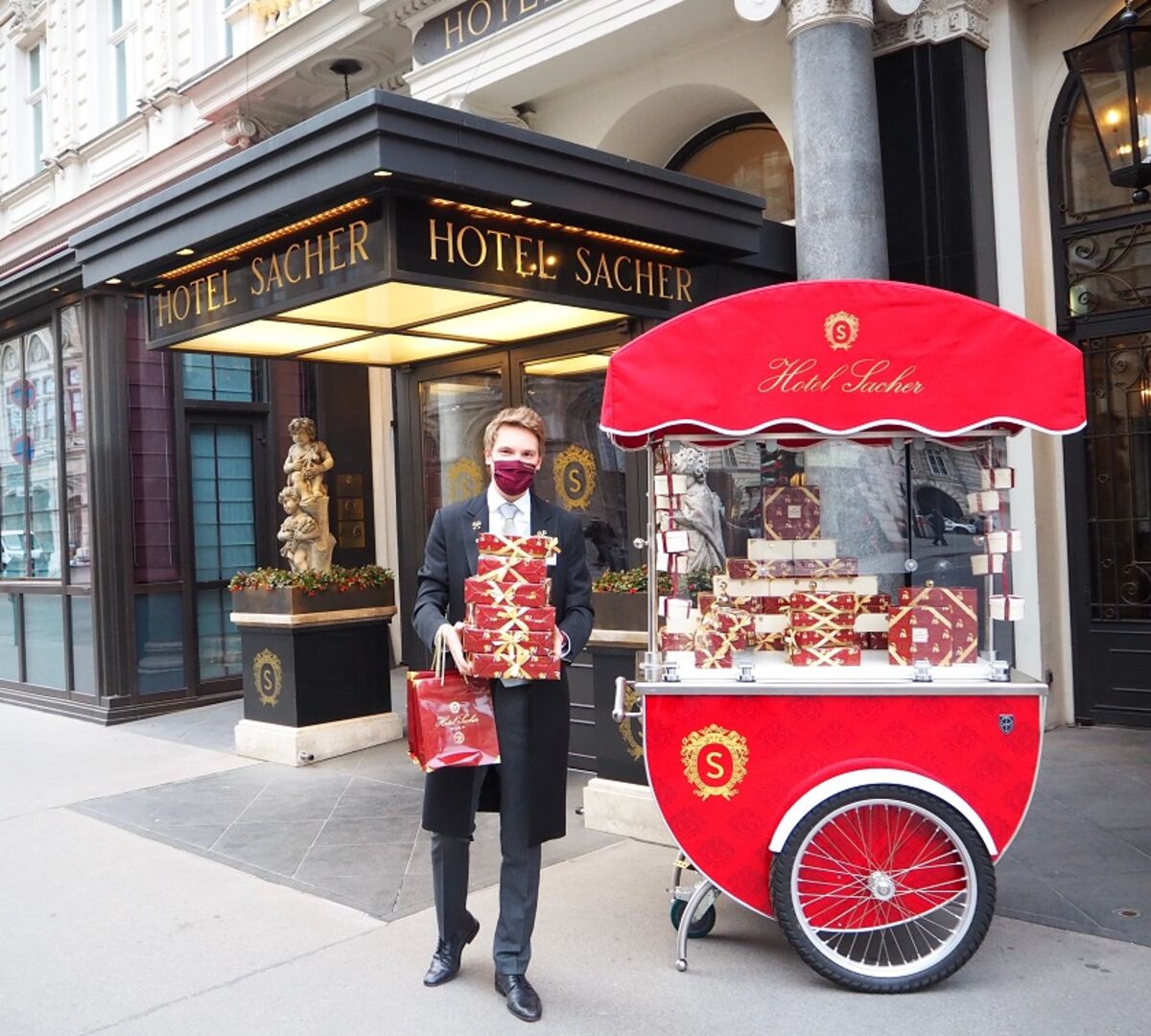 Ein Concierge mit Mund-Nasen-Schutz steht mit einem kleinem Verkaufsstand vor dem Hotel Sacher. In der Hand einen Stapel verpackter Sacher-Torten