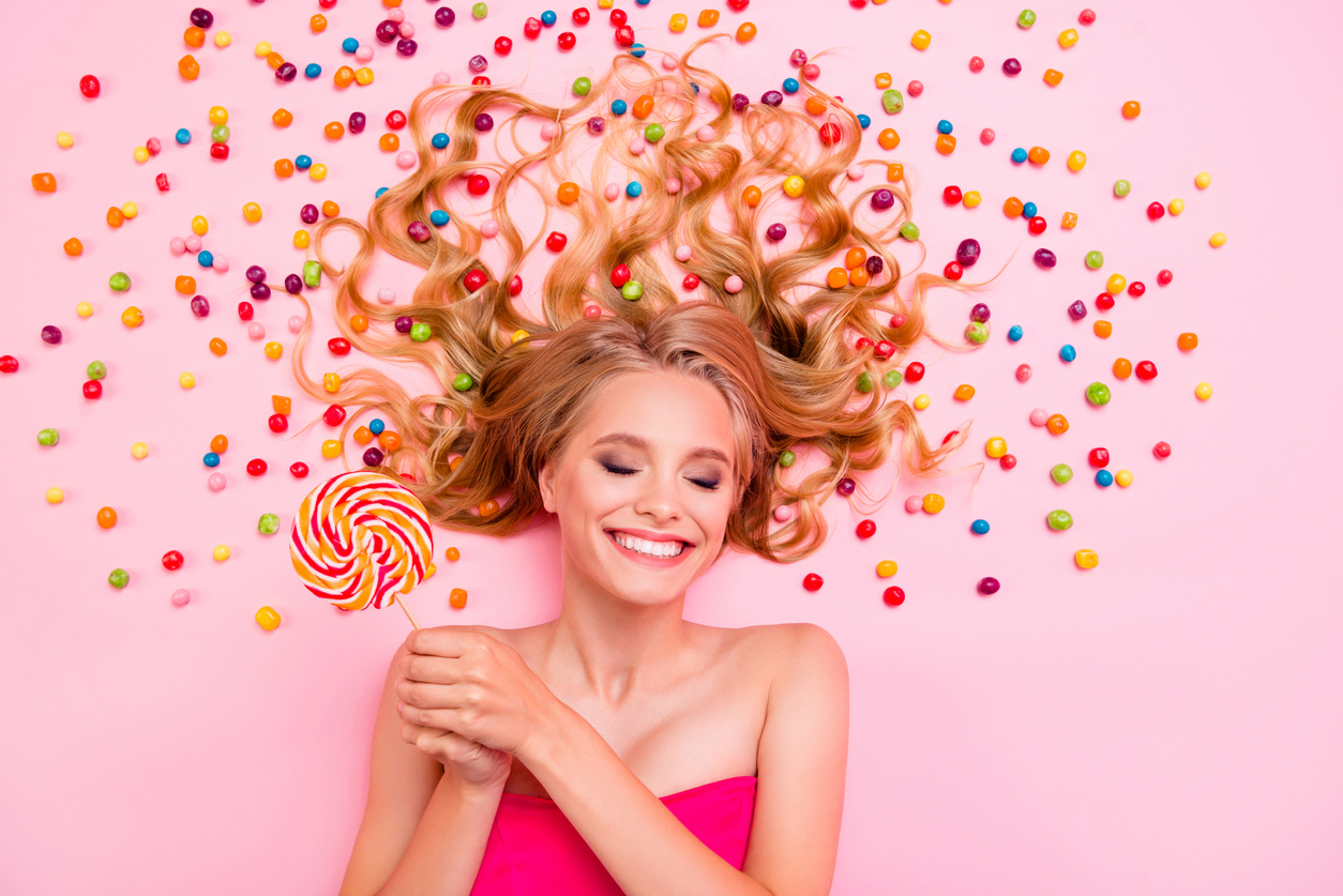 Eine junge Frau vor rosa Hintergrund mit Süßigkeiten hält einen Lolli in der Hand und freut sich.