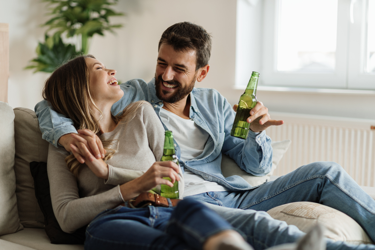 Ein Mann und eine Frau sitzen lachend Arm in Arm mit Bier auf einer Couch
