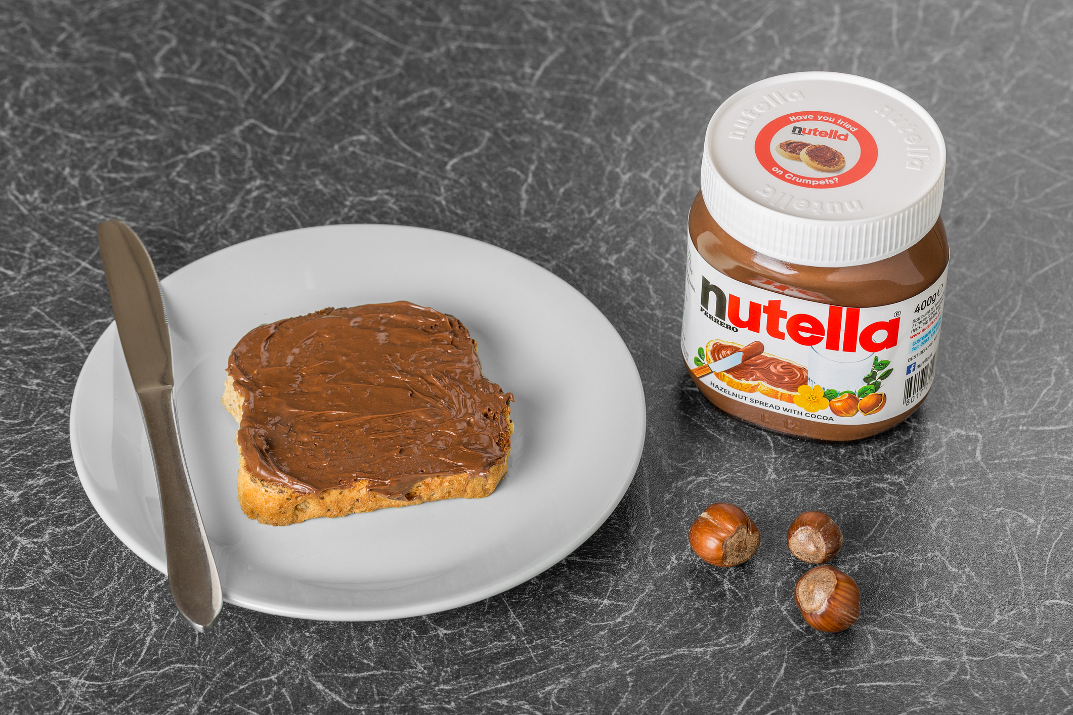 Mit Nutella beschmiertes Weißbrot neben einem Nutella-Glas und drei Haselnüssen