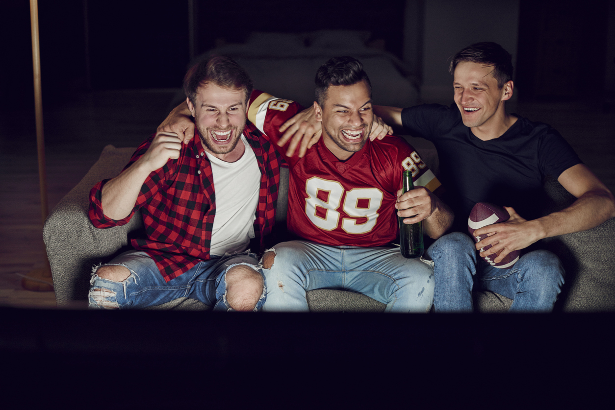 3 Männer sitzen mit einem Football und Bier auf der Couch und schauen lachend fern