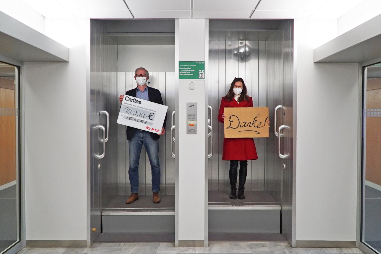 Ein Mann steht mit einem Spendenscheck in der Hand in einem Paternoster neben einer Frau, die ein Schild mit der Aufschrift &quot;Danke&quot; in Händen hält