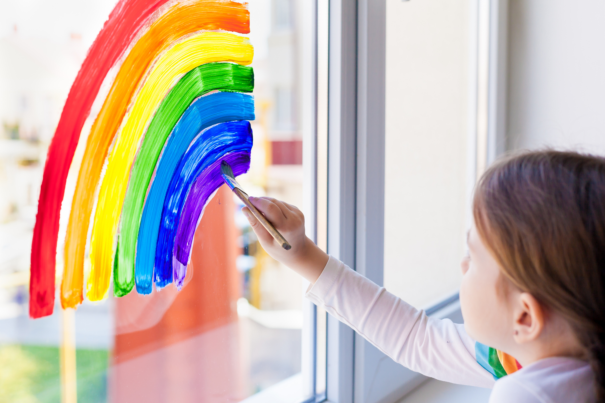 Ein kleines Mädchen malt mit einem Pinsel und Window-Color einen Regenbogen auf eine Fensterscheibe