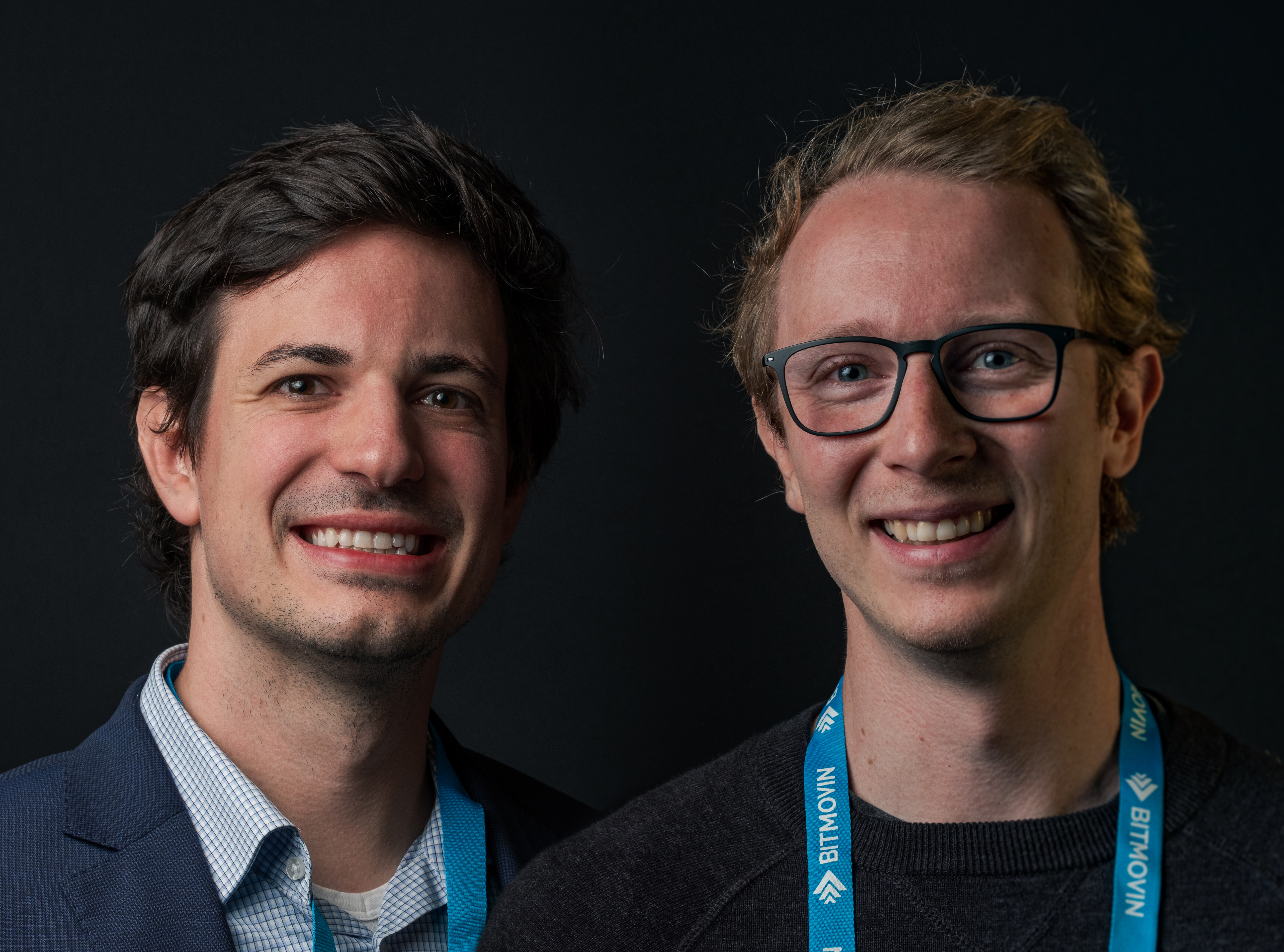 Die Bitmovin Gründer Stefan Lederer (ein junger Mann mit kurzen braunen Haaren, blauem Sakko und weißem Hemd) und Christopher Müller (ein junger Mann mit kurzen blonden Haaren, schwarzem T-Shirt und einer Brille) im Porträt