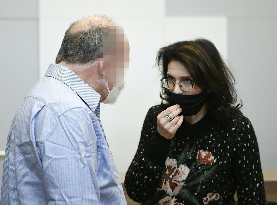 Ein Mann mit blauem Hemd steht mit dem Rücken zum Betrachter. Sein teilweise sichtbares Halbprofil ist verpixelt. Er spricht mit einer dunkelhaareigen Frau (Astrid Wagner), die eine schwarze Maske trägt.