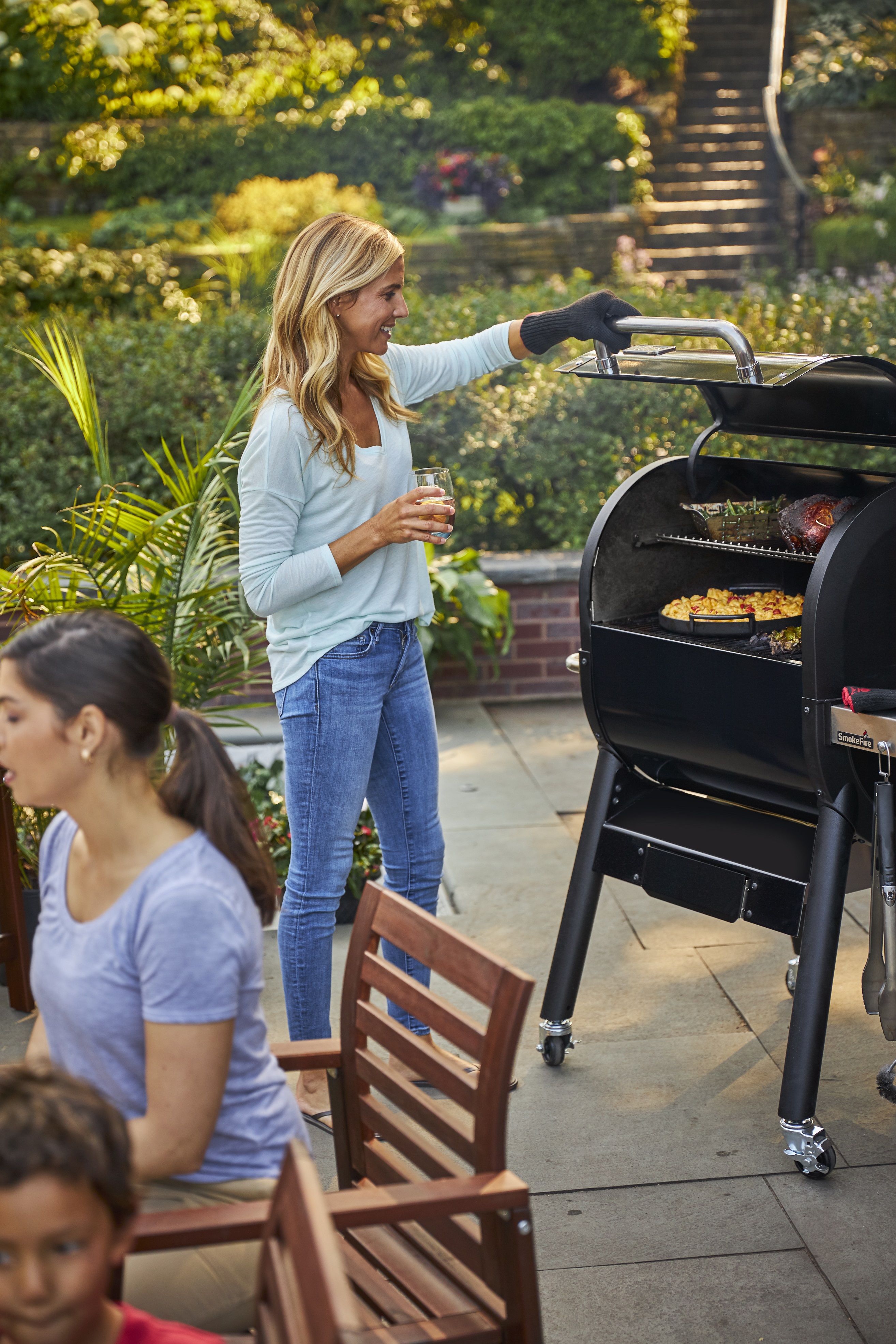 Eine junge Frau mit blonden Haaren öffnet einen Holzpelletgrill im Garten und inspiziert das Grillgut