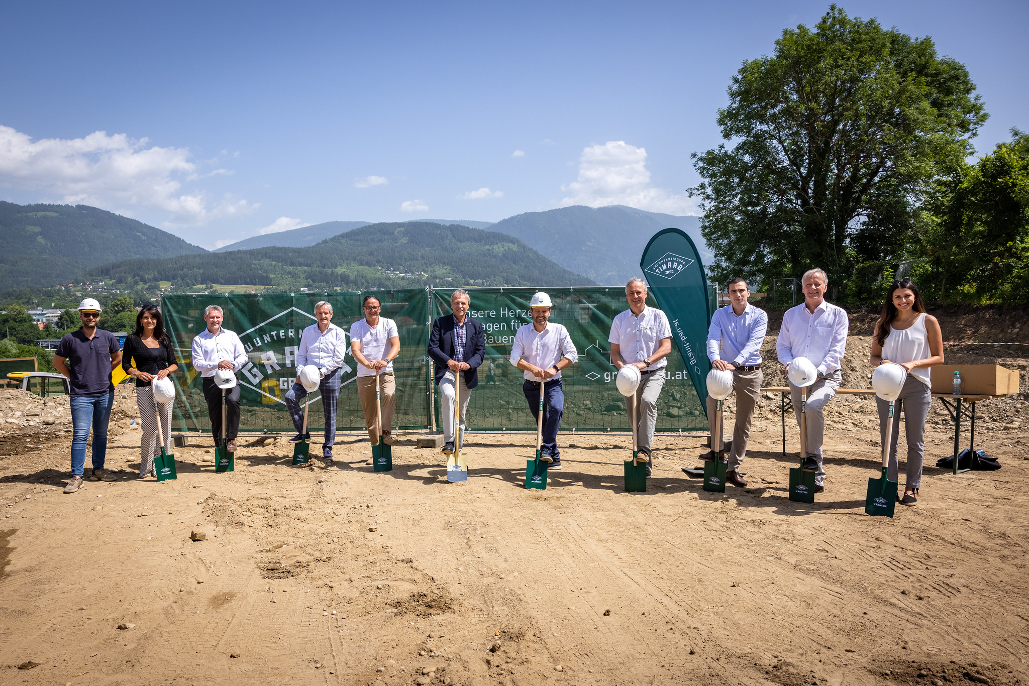Eine Gruppe von Männern und Frauen steht auf Spaten gestützt auf einem Baugrundstück mit Panoramablick