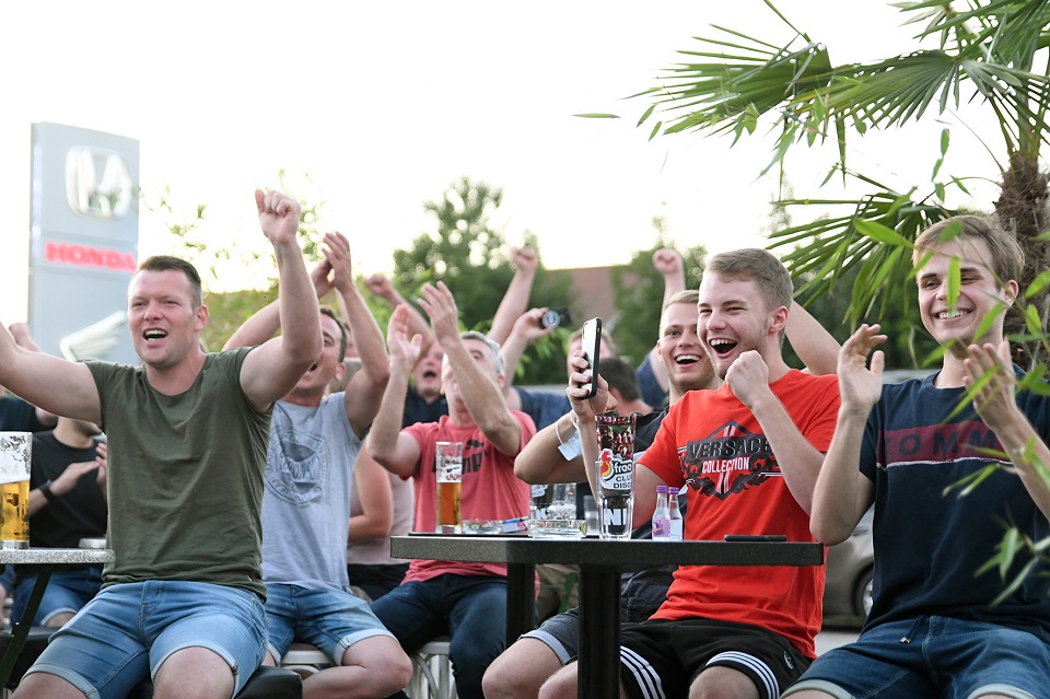 Vier junge Männer sitzen im Freien um einen Tisch mit Biergläsern, jubeln und werfen die Arme in die Luft.
