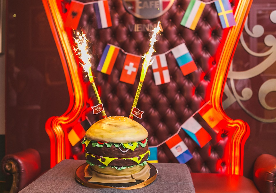 Eine Torte, die aussieht wie ein Burger mit sprühenden Wunderkerzen vor einem Ledersessel, der mit Flaggen geschmückt ist.
