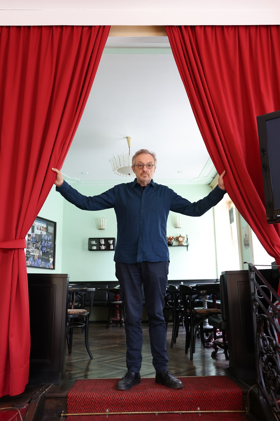 Josef Hader steht mit ausgebreiteten Händen im Rüdigerhof und hält links und rechts einen roten Vorhang. Im Hintergrund sieht man Kaffeehaus-Mobiliar.