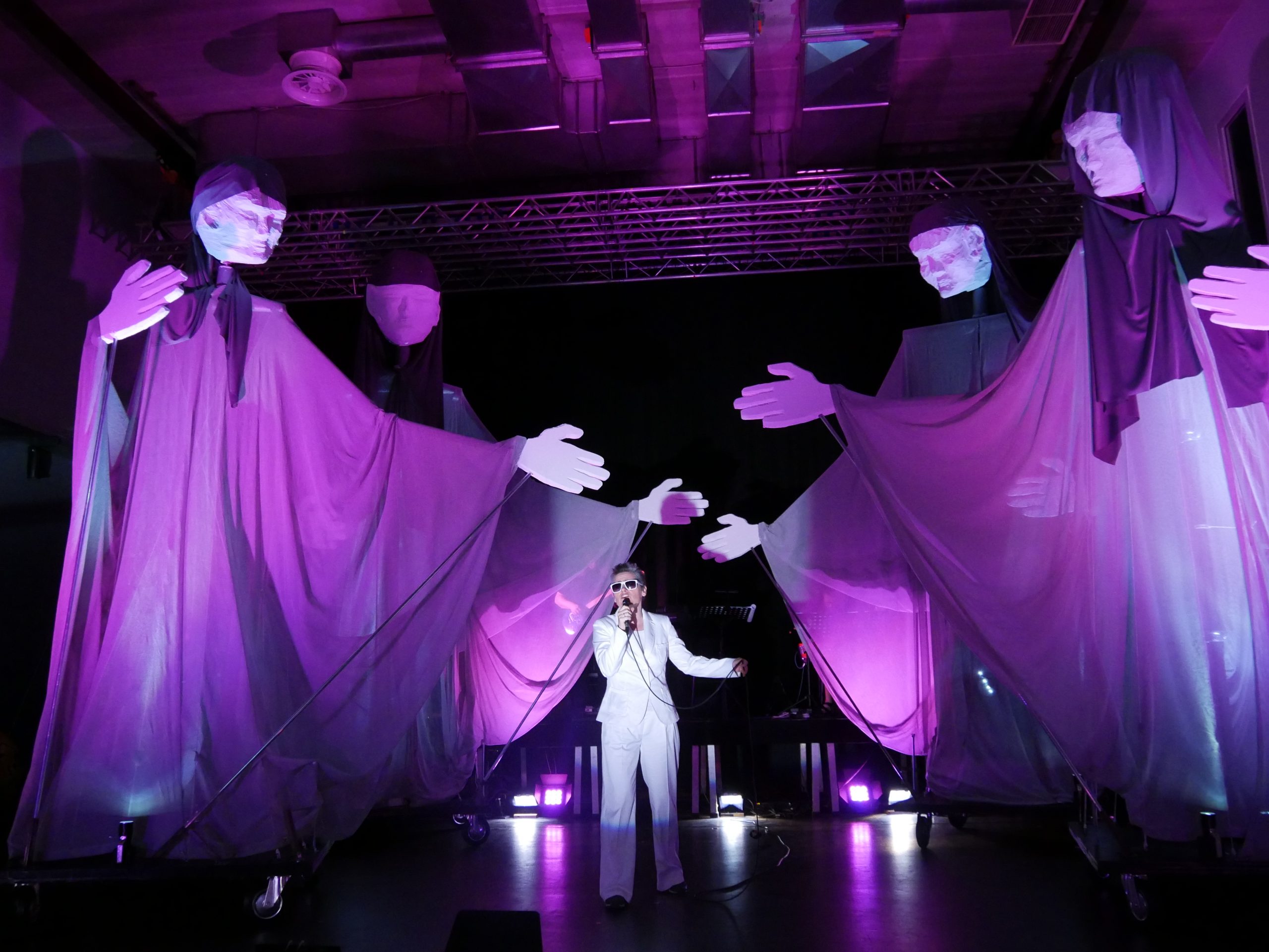 Ein violett ausgeleuchtetes Bühnenbild mit 4 überlebensgroßen Puppen und einem Sänger in ihrer Mitte