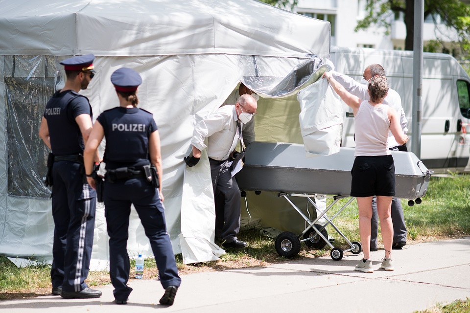 Zwei Polizisten bewachen ein Obduktionszelt. Eine Frau hält den Vorhang zur Seite. Zwei Männer mit Mundschutz schieben einen Blechsarg nach draußen.