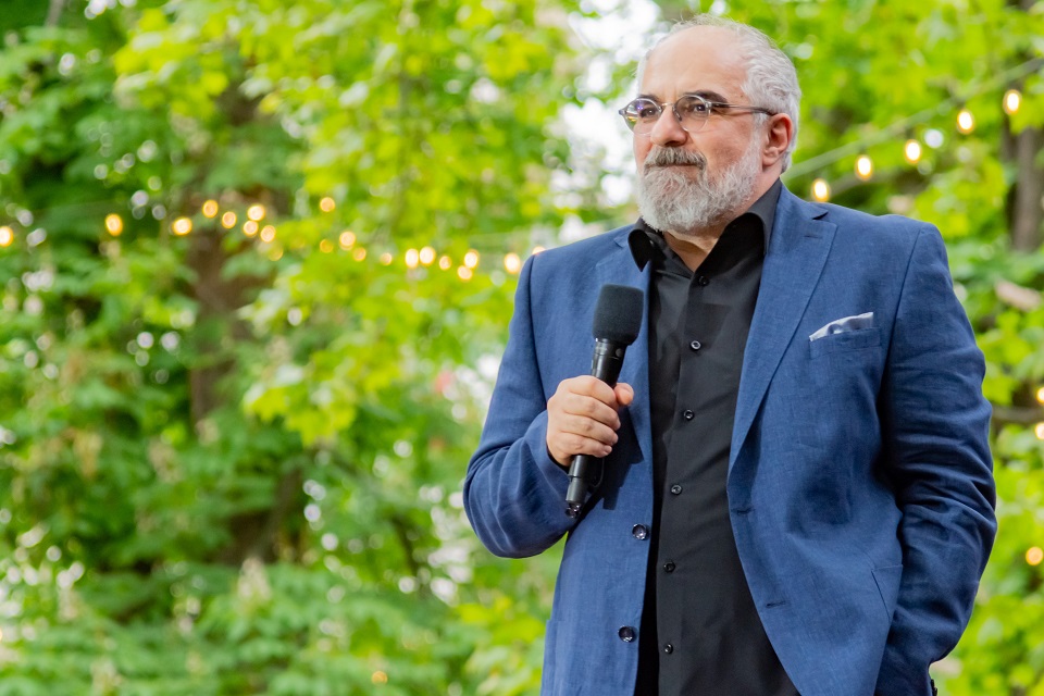 Michael Niavarani steht mit einem Mikro auf in der Hand auf einer Bühne. Man sieht ihn bis zur Hüfte. Im Hintergrund sind Bäume und eine Lichterkette zu sehen.