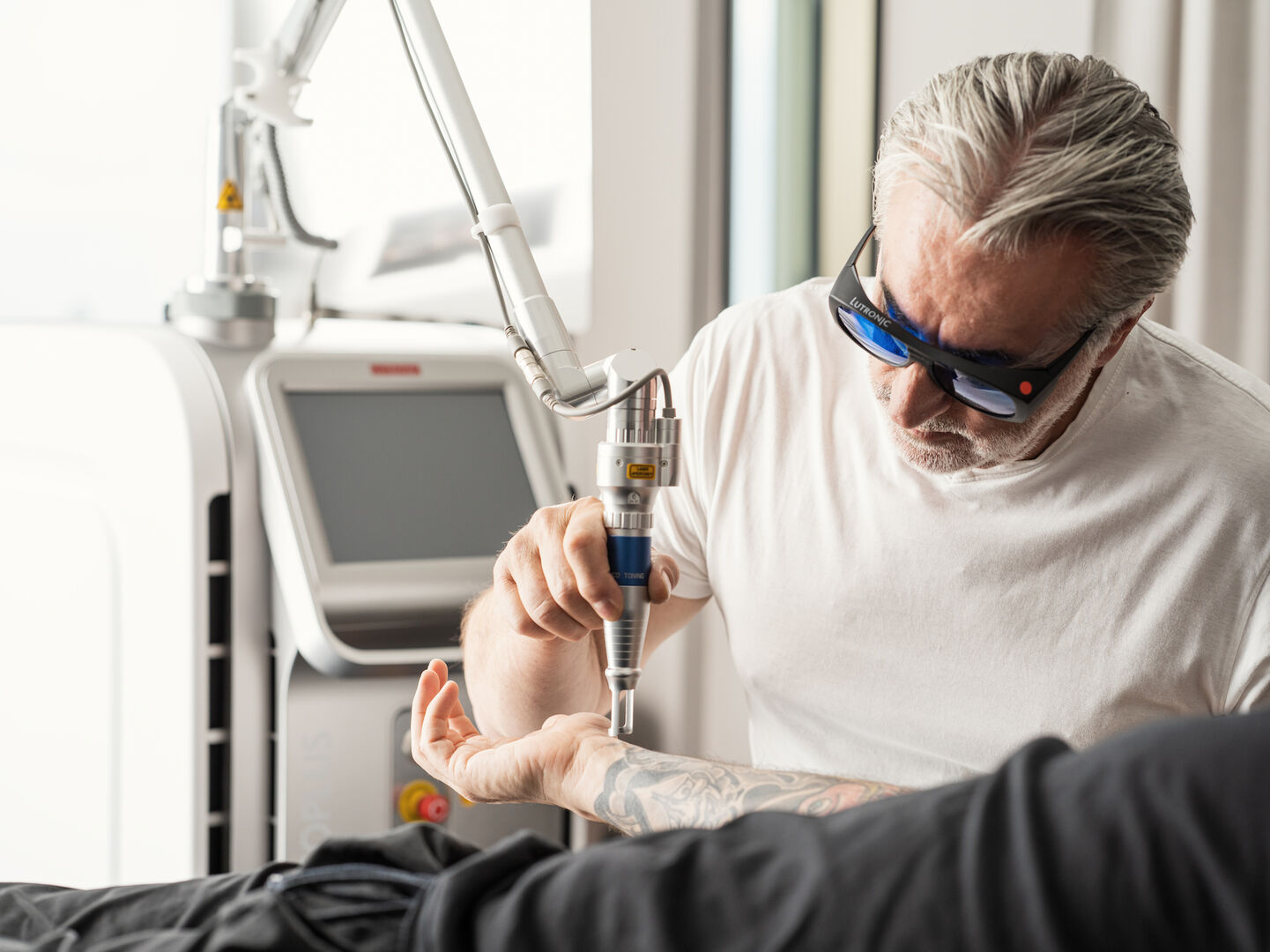 En Fachmann entfernt mittels Laser ein Tattoo am Unterarm einer liegenden Person