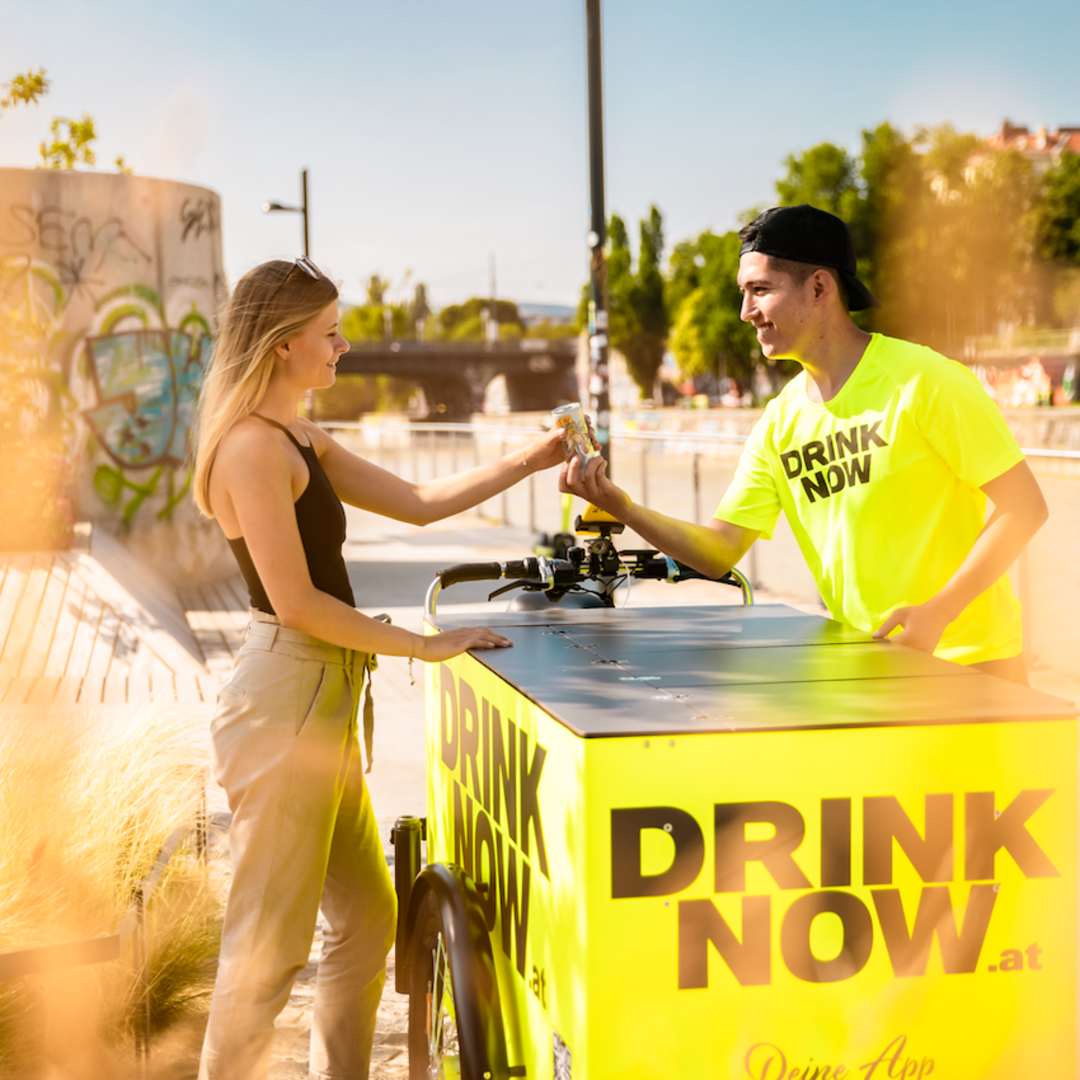 Junge Frau am neuen E-Lastenrad, junger Mann verkauft ihr ein Getränk