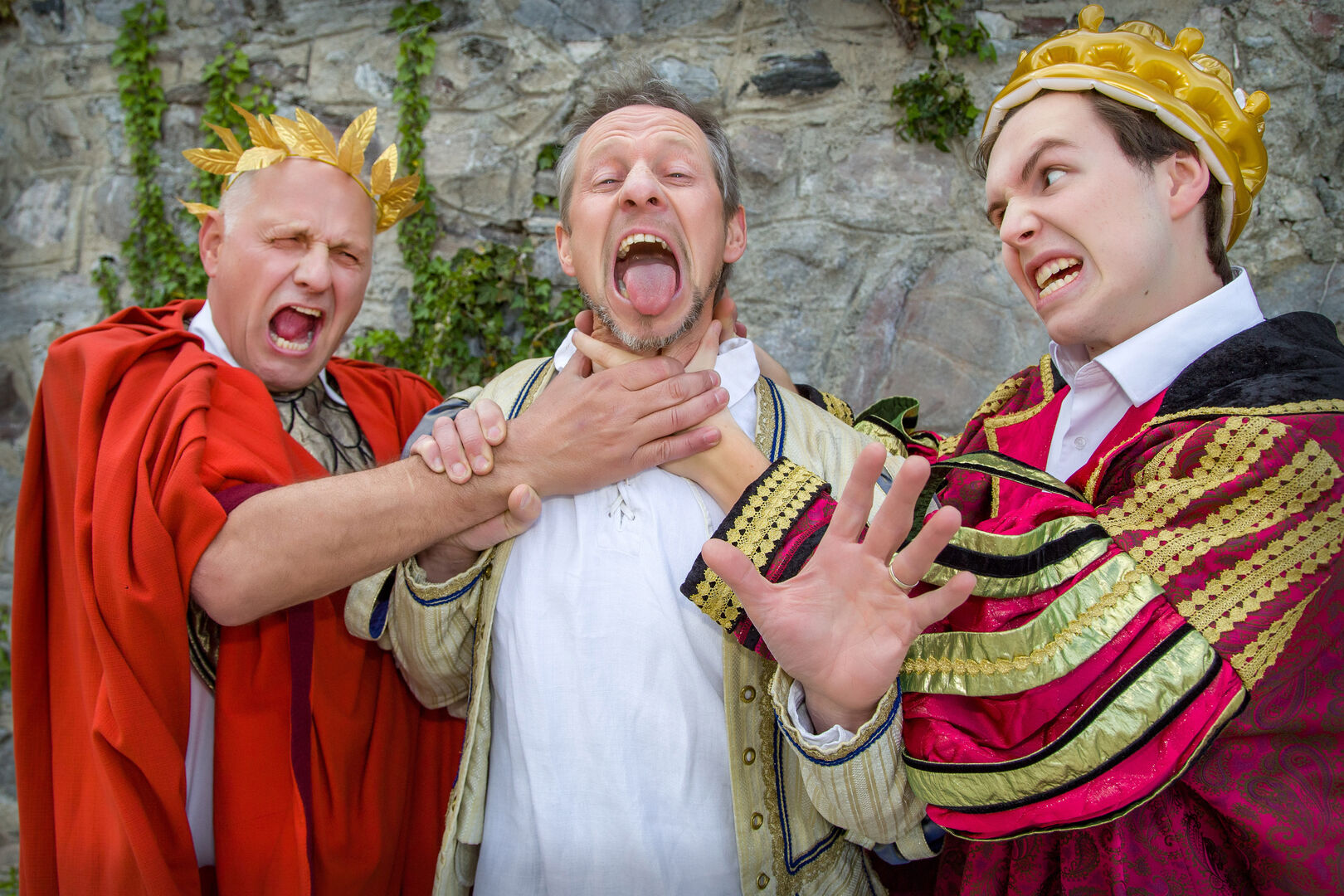 Ein Mann in einem Caeser-Kostüm und ein zweiter in der Verkleidung eines Königs würgen einen dritten im mittelalterlichen Outfit