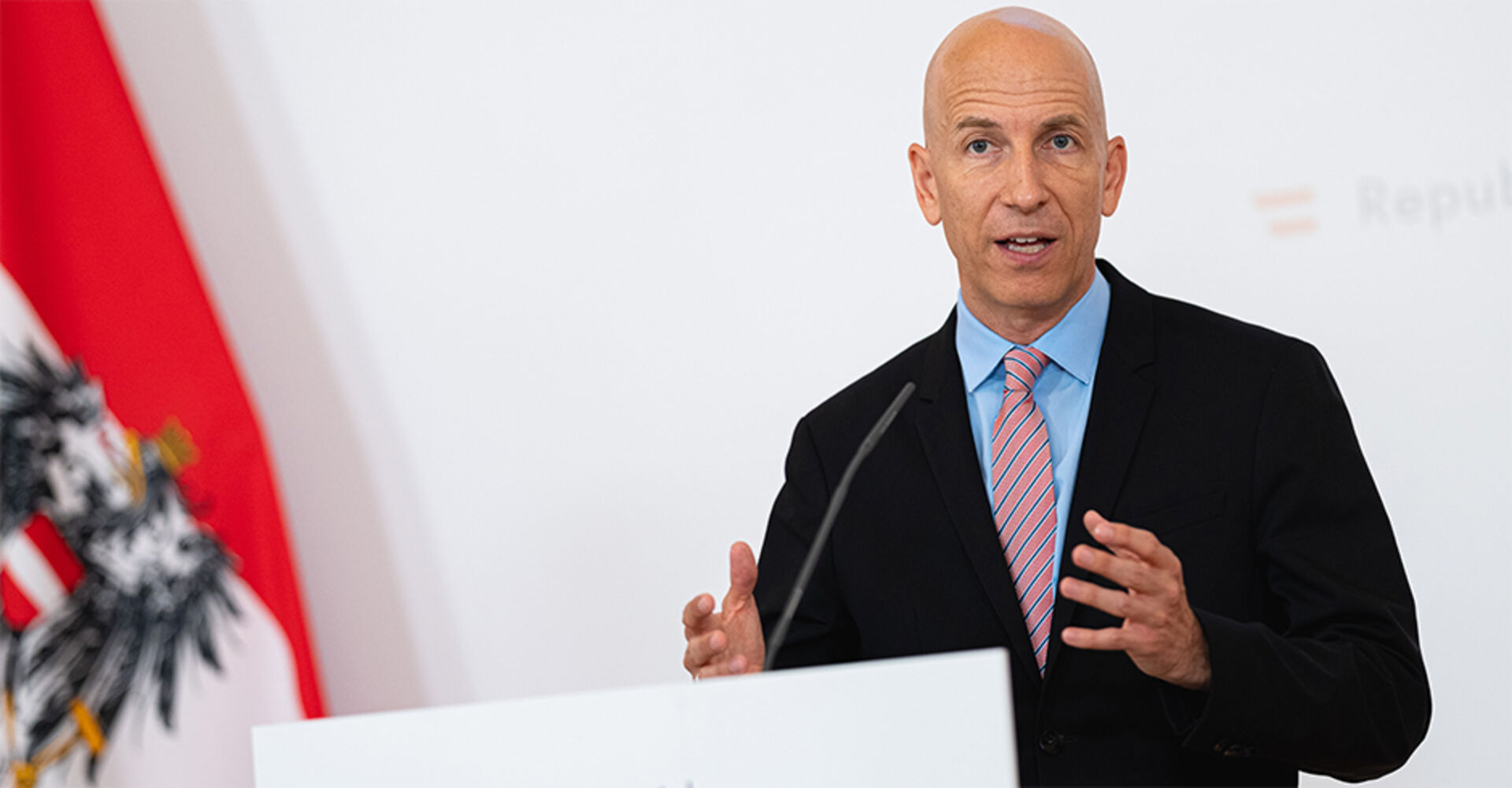 Kocher erklärt bei der Pressekonferent mit österreichischer Fahne im Hintergrund, die neuen Arbeitsmodelle