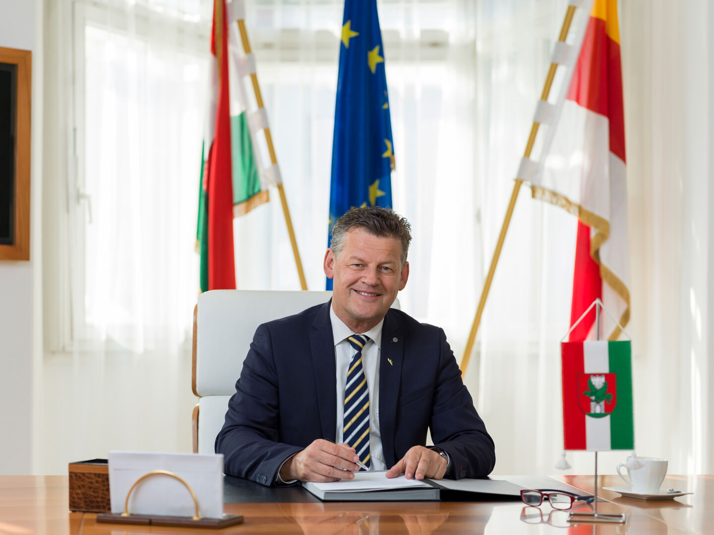Klagenfurts Bürgermeister Christian Scheider hinter seinem Schreibtisch vor einer Klagenfurt-, einer Kärnten- und einer EU-Flagge