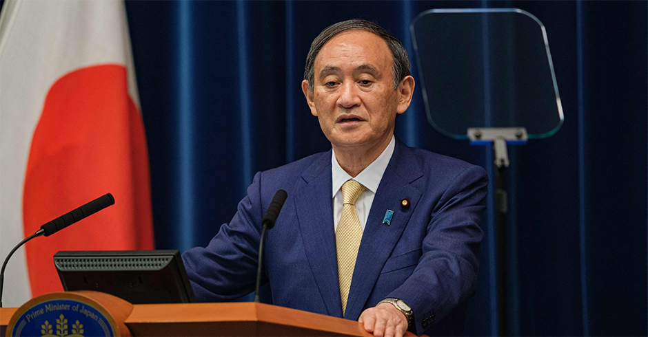 Ministerpräsident Yoshihide an einem Rednerpult vor einem dunkelblauen Vorhang und der japanischen Flagge.