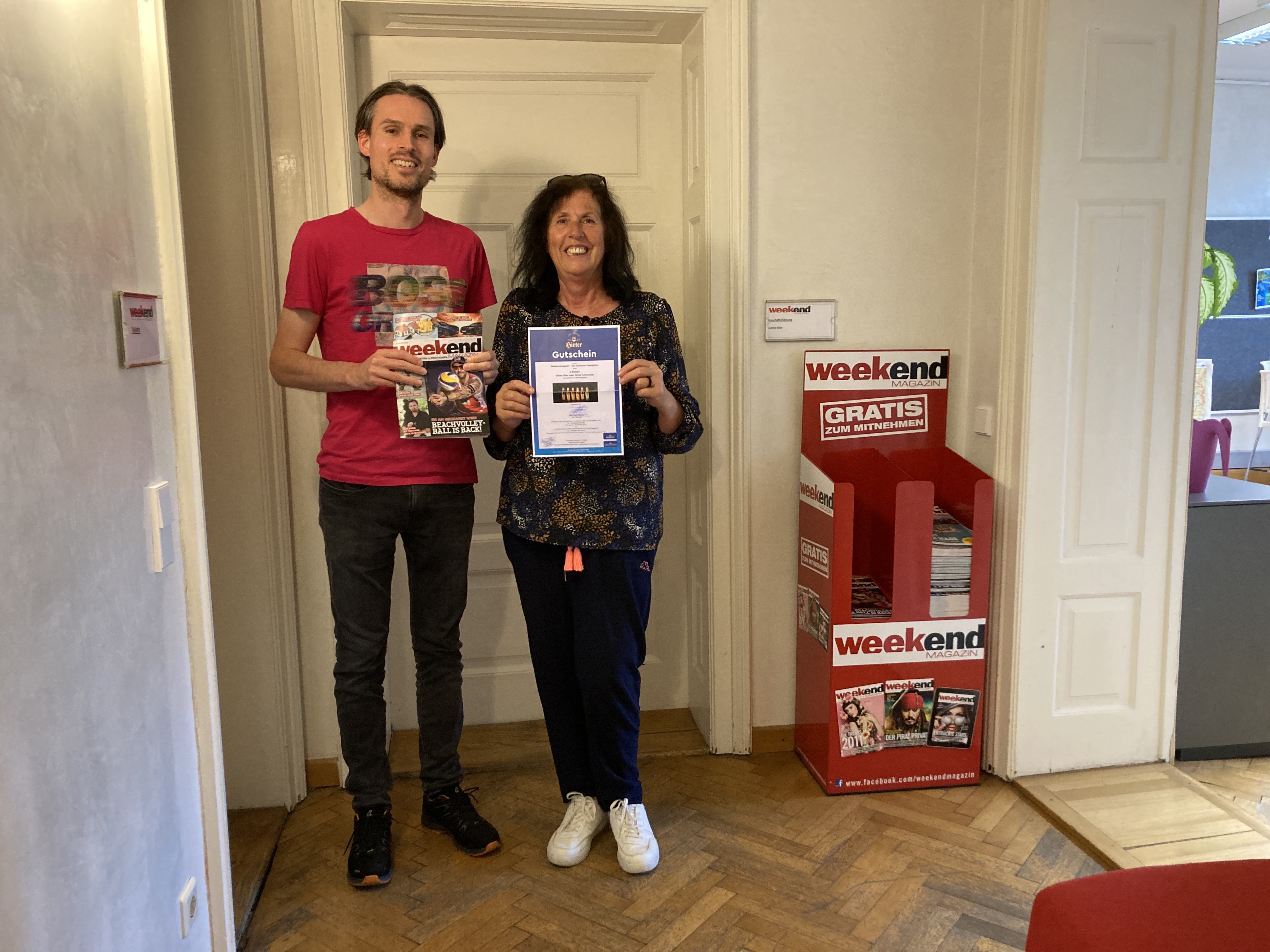 Der Weekend Chefredakteur steht in einem Büro mit einer aktuellen Ausgabe in der Hand neben der Gewinnerin des Gewinnspiels, die einen Gutschein in Händen hält