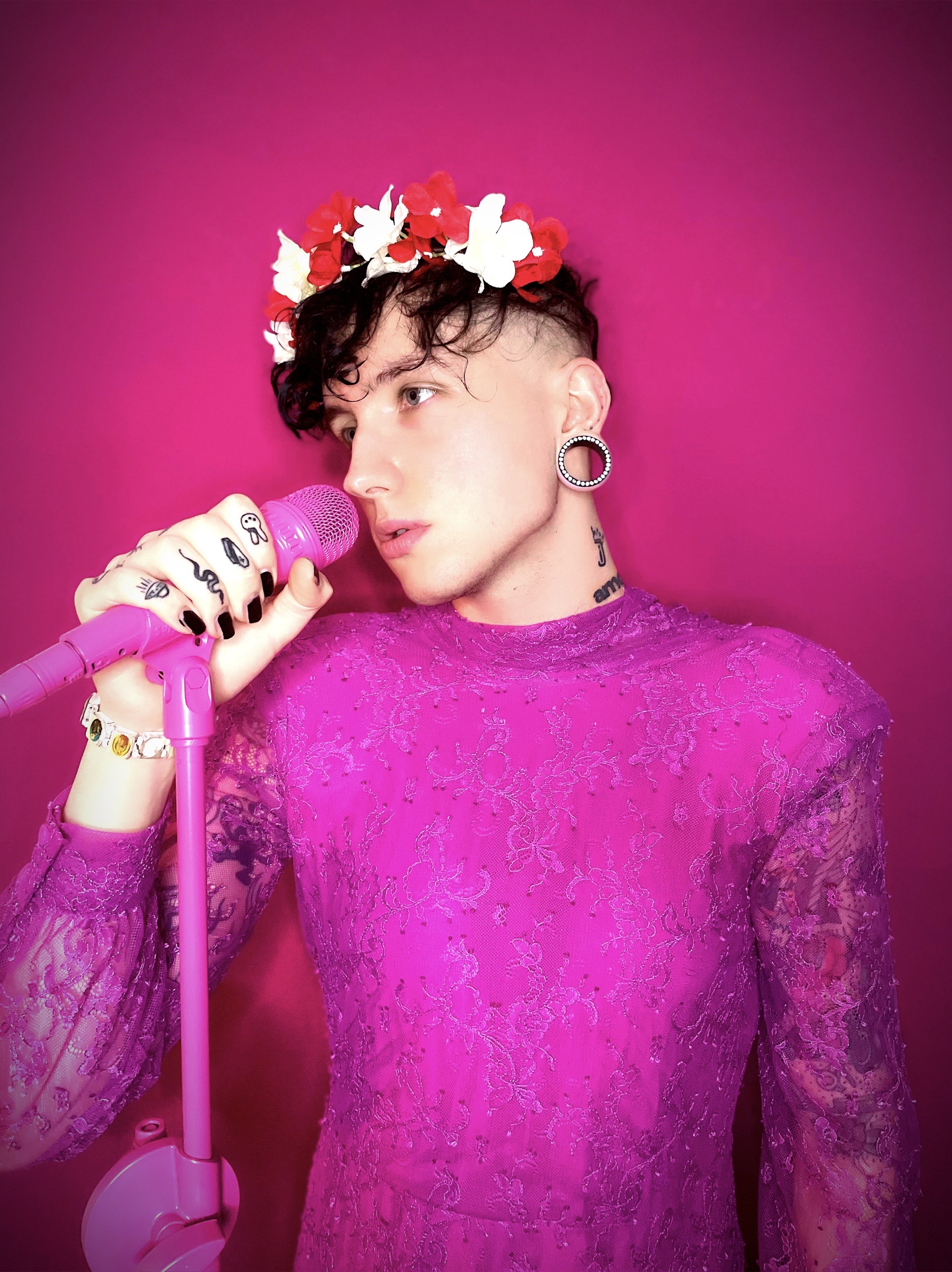 Ian Jules in einem pinken Kleid und mit einem Blumenkranz auf dem Kopf vor einem Mikrophon