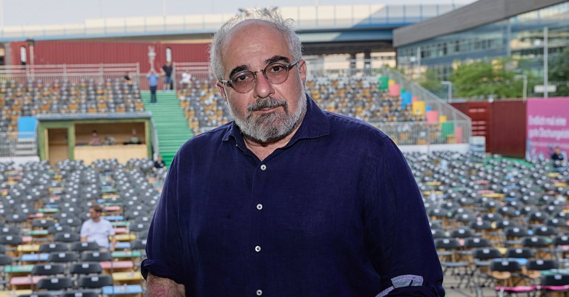 Ein Porträt von Michael Niavarani. Er steht auf der Bühne im Globe Open Air mit dem Rücken zum leeren Publikum.