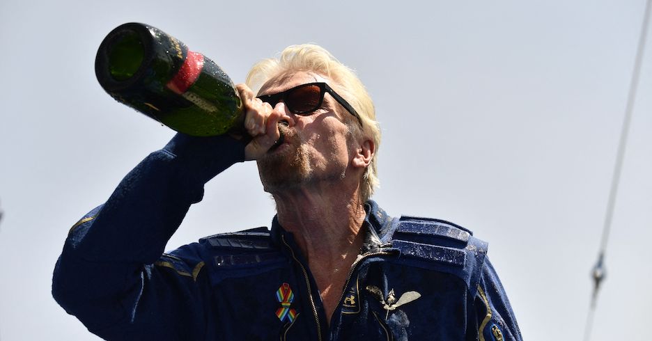 Richard Branson trägt eine Sonnenbrille und einen dunkelblauen Space-Overall. Er trinkt aus einer Flasche Champagner