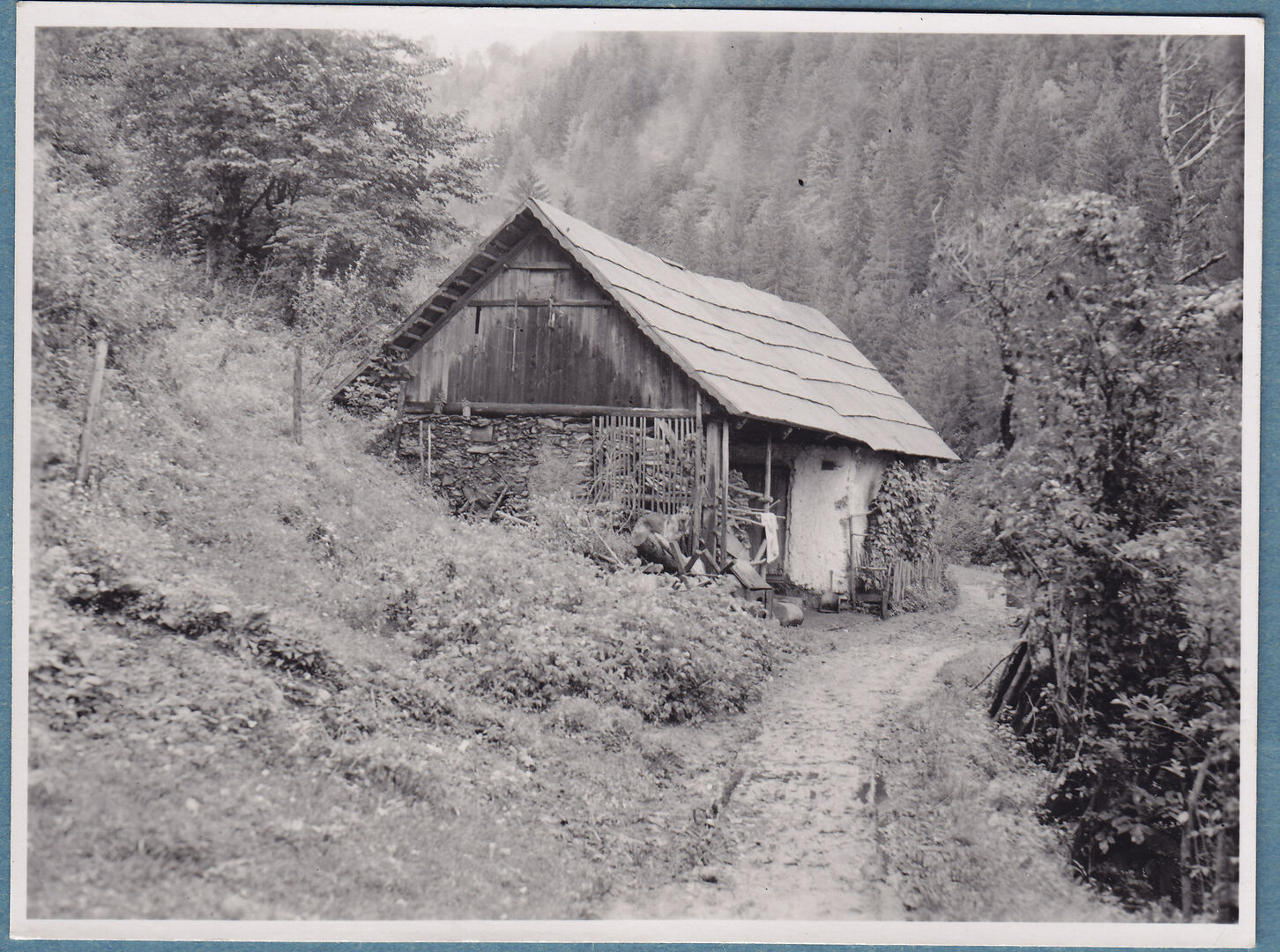 Ein Schwarz-Weiß-Foto eines einfachen kleinen Bauernhauses neben einem Waldweg