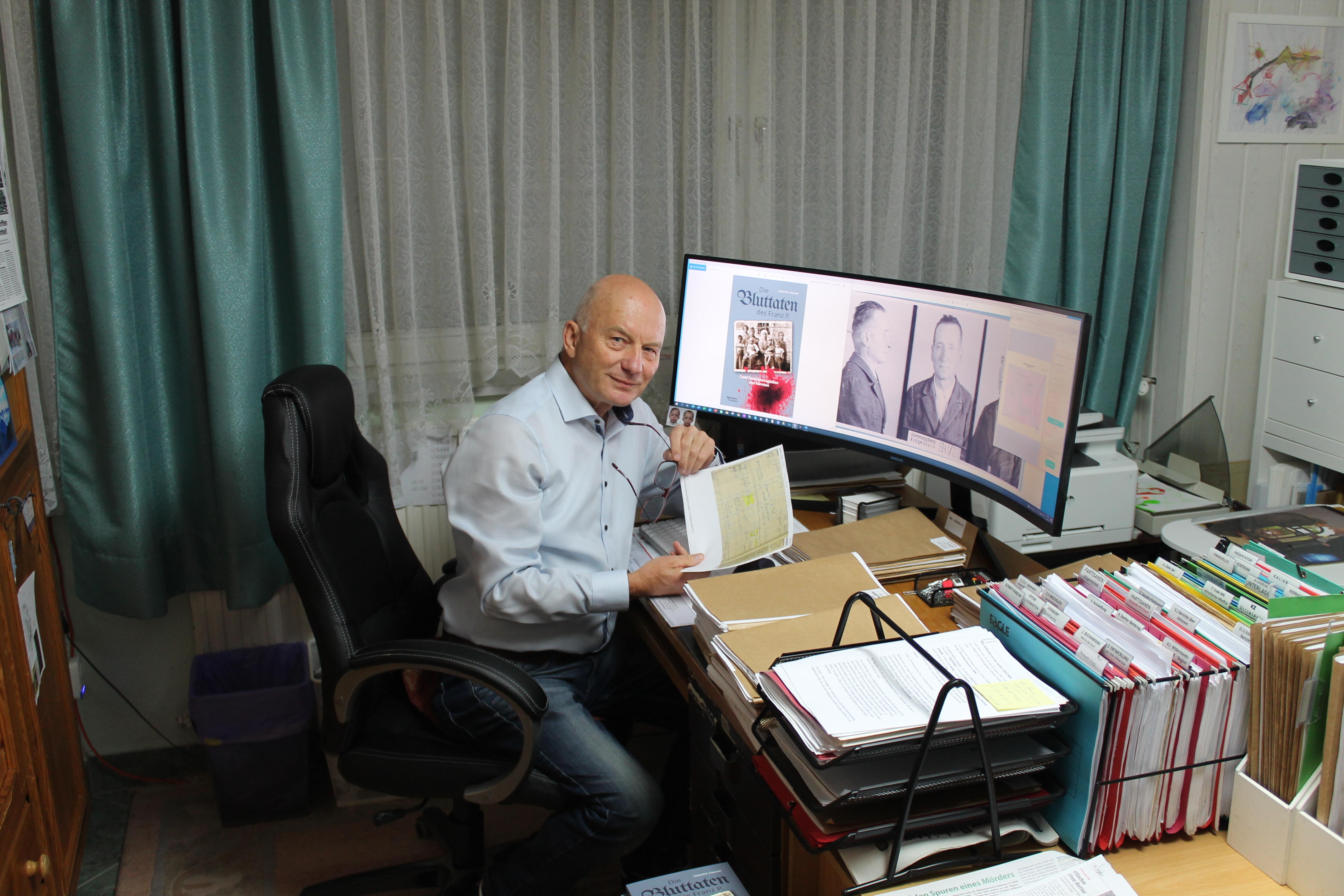 Autor Valentin Hauser sitzt vor seinem mit unzähligen historischen Akten bedeckten sowie einem großen Computerbildschirm ausgestatteten Schreibtisch