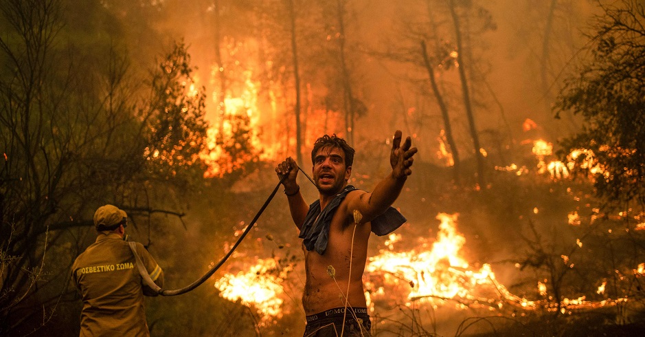 Ein Mann steht mit nacktem Oberkörper vor einem brennenden Wald. Er gestikuliert wild Richtung Kamera und hält einen Schlauch. Dieser führt zu einem Feuerwehrmann. Er versucht den Brand zu löschen.